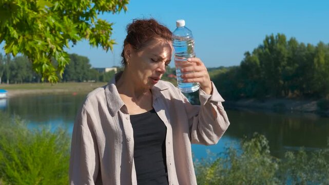 Woman drinking water by lake, feeling refreshed during hot summer day, experiencing coolness of gentle breeze while staying hydrated in scenic natural setting