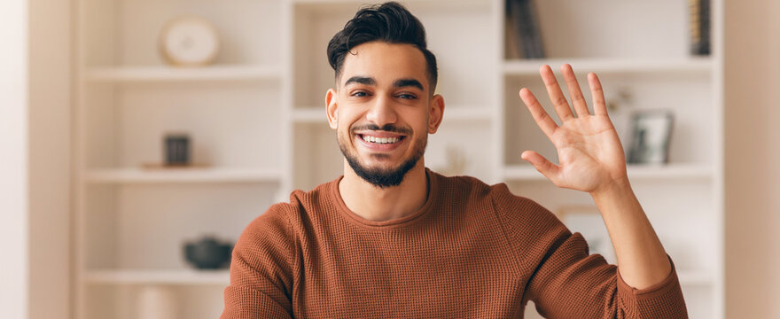 A man is sitting at a table in a well-lit room. He smiles and raises his hand in a friendly greeting. The background includes shelves with various items.