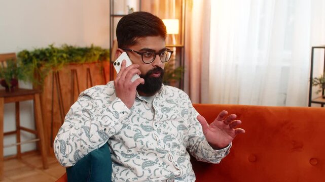Indian man at home argues on smartphone while sitting on sofa, reacting to spam call or disturbing message. Arabian guy looks frustrated, annoyed, emotionally affected, angry about mobile phone talk