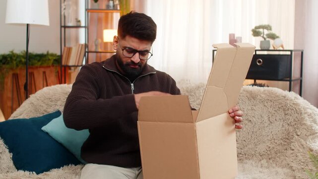 Indian man at home opens delivery box, sees incorrect item and shakes head in disappointment. Hispanic guy on sofa looks upset and frustrated by online shopping mistake and order fulfillment issue