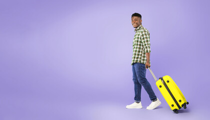 Fototapeta premium A young man is walking while pulling a bright yellow suitcase. He is smiling and dressed in checkered clothing. The background is a solid purple color, suggesting a fun and vibrant atmosphere.