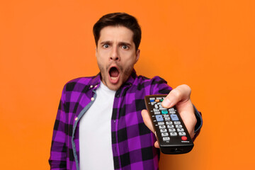 A man stands with a shocked expression while pointing a remote control toward the camera. His shirt is white under a checkered purple and black jacket. The background is a bright orange color. © Prostock-studio