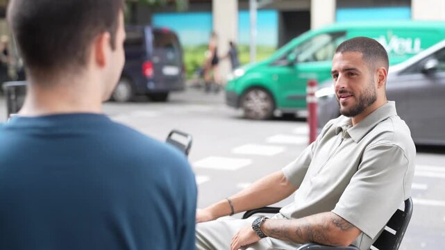 Man sits in wheelchair, smiling gently. Another man faces him, attentive and engaged. Background shows urban street with vehicles passing. Scene captures quiet, meaningful conversation outdoors