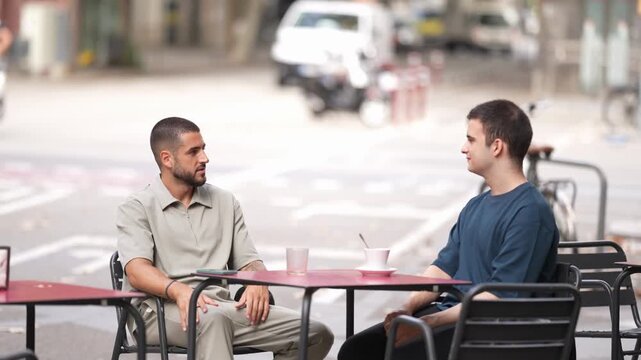 Two men sit at a street cafe table. They sip drinks while chatting casually. Background traffic moves softly behind them. Urban setting feels relaxed and informal