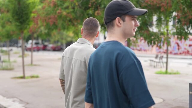 Two men walk side by side through a city park. One wears a cap, the other a light shirt. Trees line the path behind them. Mural adds color to a distant wall