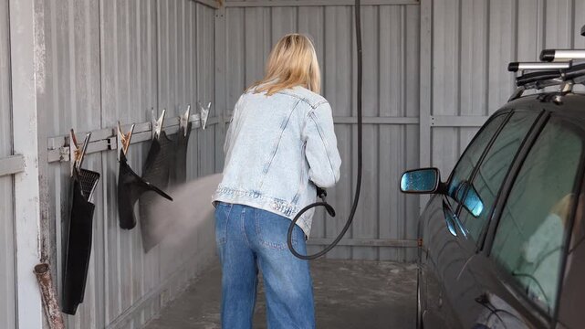 Blonde woman wearing a denim jacket carefully cleaning floor mats and the side of her dark vehicle with a powerful jet of water at a self-service car wash station in an urban area