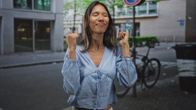 Thai woman in blue blouse raises fists with bare hands visible while smiling near a bicycle against an urban building on a street; triumph.