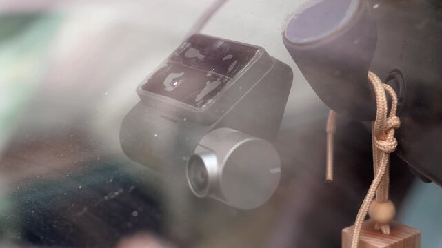 Modern car dashcam mounted on a dirty windshield recording the road for safety and surveillance purposes. An air freshener is hanging next to the digital video recorder capturing the commute