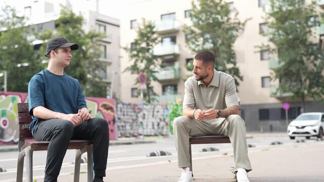 Two men sit on a bench, chatting casually. They wear casual clothes and show relaxed expressions. Trees and buildings form a quiet urban background