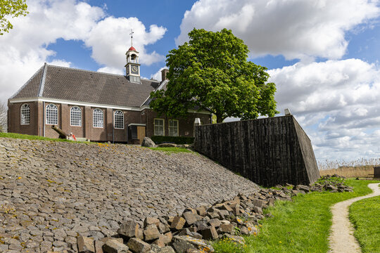 View of the historic church of Schokland with its bell tower and a large green tree behind a stone embankment under a cloudy sky in Schokland, Flevoland, Netherlands.