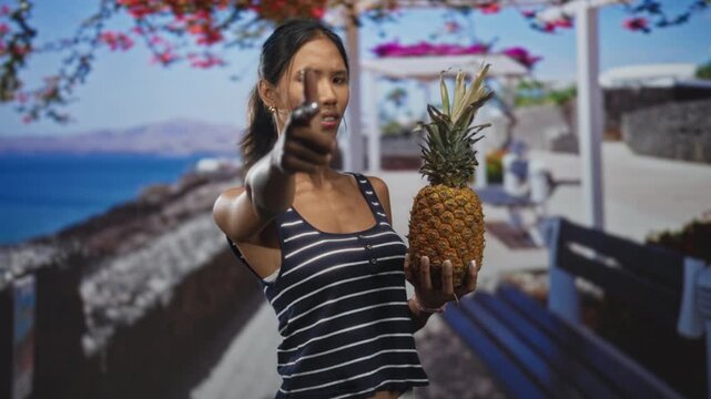 Thai woman in striped tanktop holds ripe pineapple with left hand and points finger to camera in studio; playful summer fun.