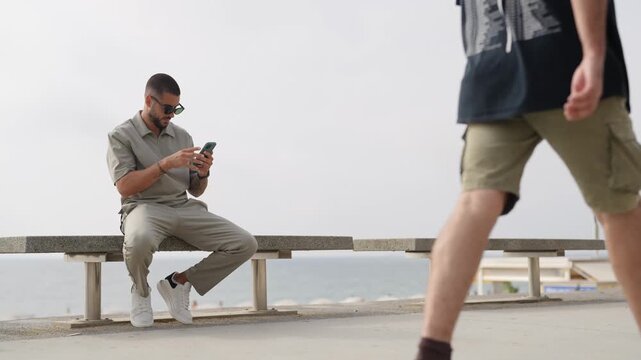 Man sits bench, engrossed in phone. Another walks past, ignoring his screen. Ocean looms behind, calm and vast. Ideal for travel or urban lifestyle scenes. No animals or plants in frame