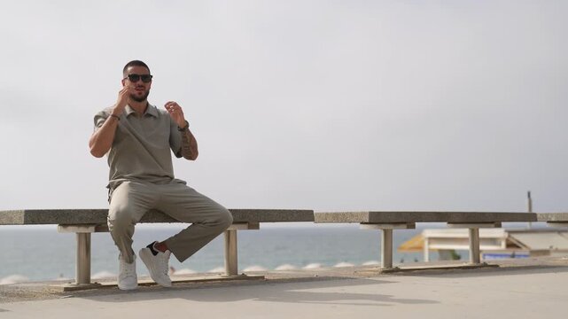 Man relaxes on bench by seaside. He adjusts sunglasses, smiles subtly. Ocean breeze drifts behind him. Perfect for tranquil beach scenes. Solemn mood, calm coastal vibe