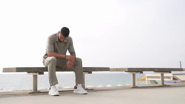 Man sits alone on bench, head bowed, gloomy, tired. He looks up slowly, expression shifts to quiet contemplation. Ocean stretches behind him, empty, indifferent, vast. Footage evokes solitude, pause