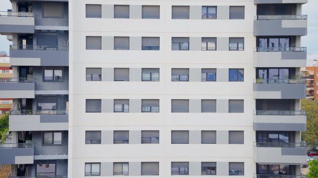 Symmetric facade of a modern residential building with white exterior walls, gray balconies with glass railings and roller shutters across multiple floors