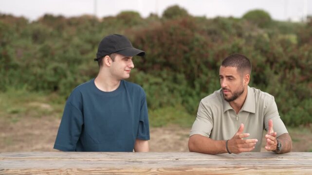 Two men converse at a wooden table outdoors. One listens attentively, the other speaks earnestly. Natural light bathes the scene, enhancing calm tones. Lush greenery frames their relaxed