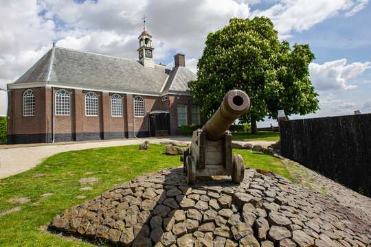 View of the historic Middelbuurt church and a vintage cannon on a stone mound under a cloudy sky in Schokland, Flevoland, Netherlands.