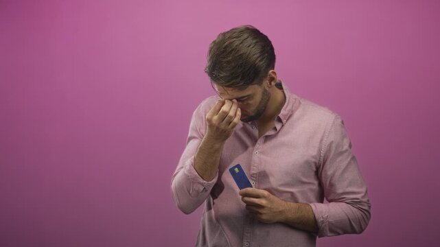 Bearded hispanic man pinching the bridge of his nose holds a blue creditcard against a solid pink backdrop in a studio; financial stress.