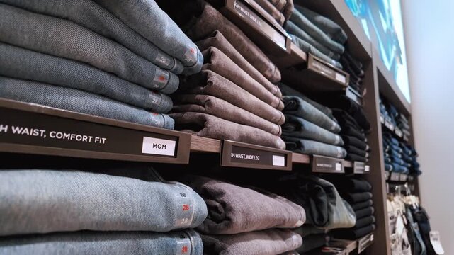 Shelves stacked high with a variety of folded denim jeans in a retail clothing store, showcasing different styles, sizes, and colors available for purchase by customers shopping in the background