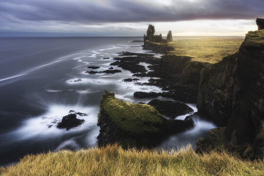 View of Londrangar basalt cliffs and the dramatic coastline with crashing waves under a cloudy sky at sunset in Hellnar, Snaefellsbaer, Iceland.