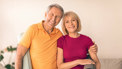 A couple stands together in a modern living room. They smile at the camera while embracing each other. The setting is bright and welcoming, suggesting a moment of happiness. © Prostock-studio