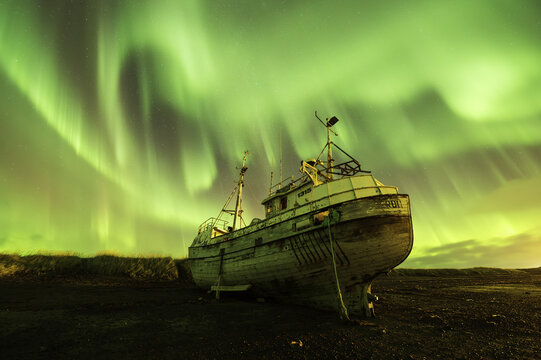 View of an old grounded wooden fishing boat under the vibrant green Northern Lights dancing across the starry night sky Olafsvik, Snaefellsbaer, Iceland.
