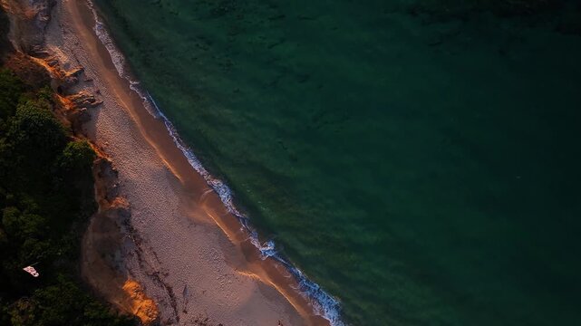 Aerial view of the golden sandy shoreline and gentle turquoise waves crashing on the beach at sunset in Costa Paradiso, Sardegna, Italy.