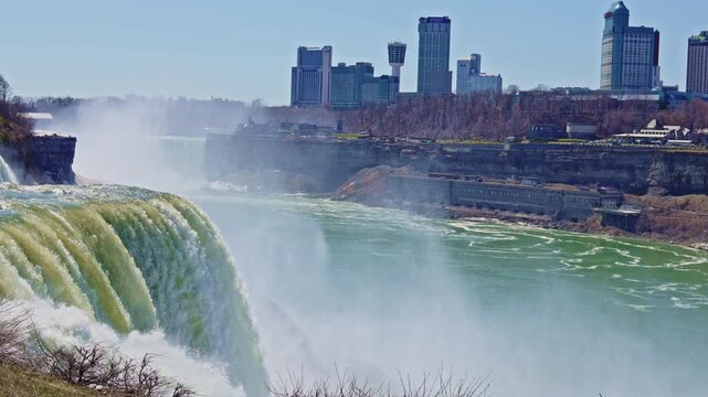 Water rushes over a large waterfall while people look on from a nearby area. Tall buildings stand in the background on a clear day next to the flowing river.