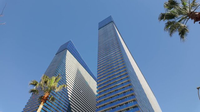 Low-angle view of two contemporary glass skyscrapers with distinctive architectural design, tracking shot reveals tall palm trees against a clear blue sky, showing a modern financial district
