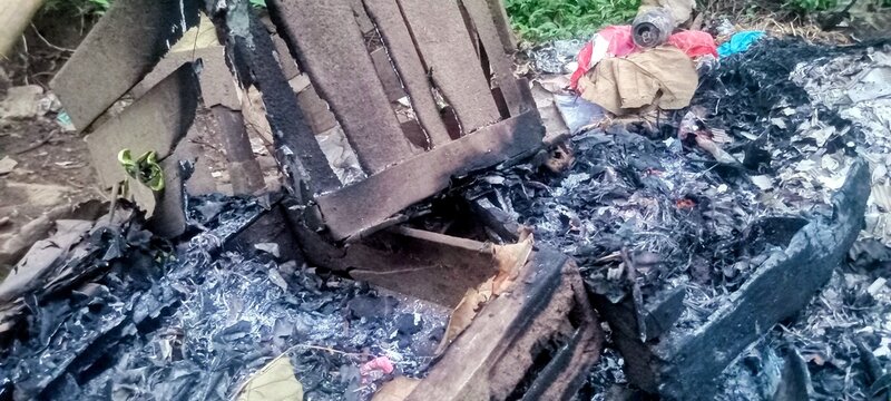 The aftermath of a fire reveals a pile of grey ashes, charred wood, and burnt debris. Fragments of cardboard, leaves, and blackened remnants are scattered across the ground. A small red ember still gl