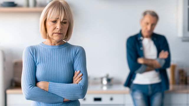 A man and woman stand in a modern kitchen, displaying signs of tension. The woman looks concerned while the man appears distant with crossed arms. They seem to be facing a conflict.