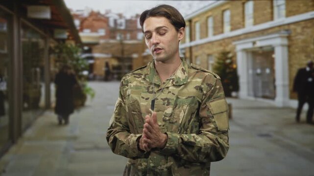 Soldier in camouflage with visible dogtags clasping hands on cobbled street in front of brick building, facing camera; duty pride.