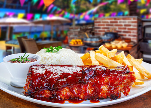 Grilled BBQ pork ribs with barbecue sauce, served with white rice and crispy french fries on a white plate, Festa Junina outdoor background
