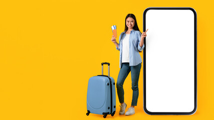 A woman stands next to a large smartphone while holding a travel ticket. She smiles and points with one finger. A suitcase is placed beside her. The background is bright yellow. © Prostock-studio