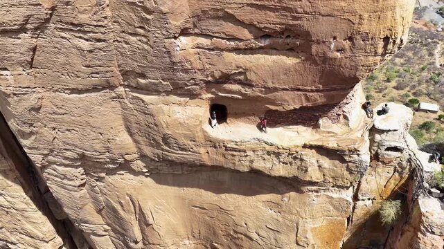 Aerial view of the Abune Yemata rock-hewn church entrance on a steep sandstone cliff with people standing on the narrow ledge in Megab, Tigray, Ethiopia.