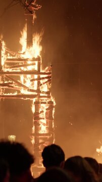 Tall lattice falla wooden framework fully engulfed in fire during Las Fallas La Crema celebration in Valencia, sparks and embers flying through orange sky as crowd silhouettes watch from below