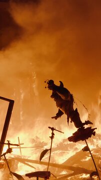 Low angle view of a burning Las Fallas falla sculpture figure silhouetted against intense orange fire glow during La Crema in Valencia, collapsing wooden debris surrounding the structure