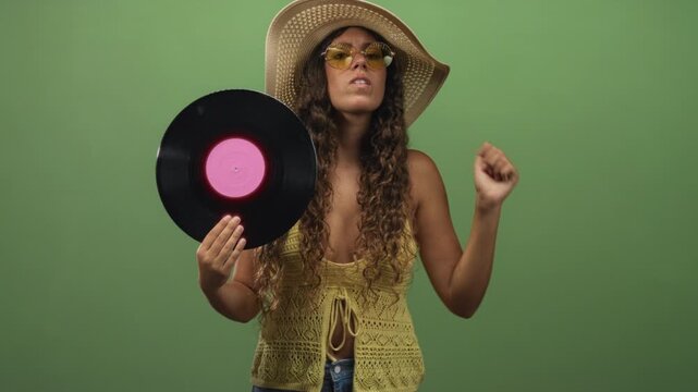 Woman holding vinyl record in studio, wearing sunhat and yellow sunglasses with hand raised in a dance gesture; playful retro music.