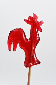 A close-up studio photograph of a glossy red rooster-shaped lollipop on a stick