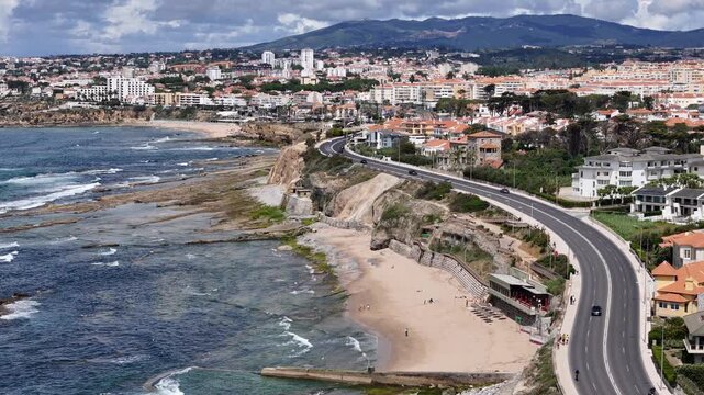 Aerial view of the scenic coastal road winding along the beach and rocky shoreline with urban buildings and distant hills in Parede, Lisbon, Portugal.