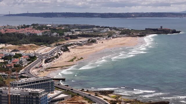 Aerial view of Carcavelos beach with waves crashing on the shore, coastal road, and Sao Juliao da Barra Fort in Lisbon, Portugal.