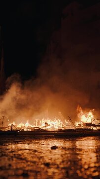 Ground level view of burning Las Fallas falla debris during La Crema in Valencia, flames and glowing embers reflected in wet cobblestone pavement with dramatic smoke rising into dark sky