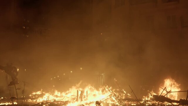 Wide view of burning collapsed falla debris spread across a Valencia street after La Crema, scattered flames and embers under thick smoke haze with orange atmospheric glow