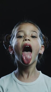 Young girl with space buns hairstyle opening mouth and sticking out tongue against black background