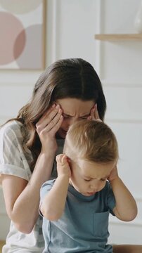 Stressed young woman and toddler boy in blue t-shirt with hands over ears, medium shot interior