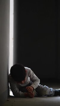 Young boy sitting on floor against wall in dark interior with backlighting medium shot