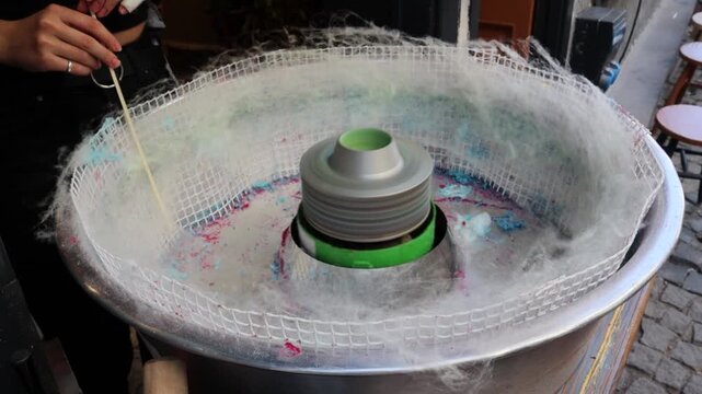 Close view video of cotton candy machine spinning fine sugar strands while a hand shapes the sweet on a stick, showing street dessert preparation, confectionery craft, snack service, and fair food.