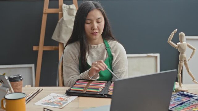 Woman in green apron selecting a colored pencil from an open case while referencing a laptop and sketch on a table in an art studio; creative concentration.