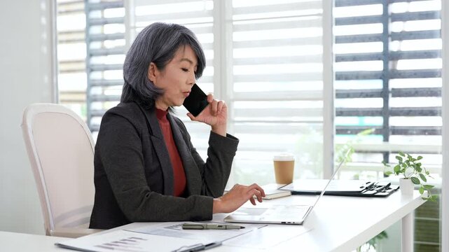 Senior businesswoman having call on smartphone with business partner.  Entrepreneur talking by mobile cellphone while working on laptop computer in office.