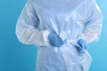 Fototapeta premium Medical worker tying disposable isolation gown on light blue background, closeup